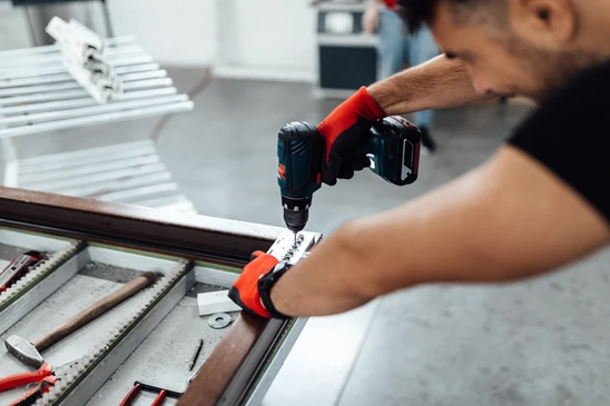 Artisan menuisier en tenue professionnelle discutant avec un client devant une fenêtre ou porte d'entrée à son domicile, montrant plans ou documents techniques, illustrant le diagnostic et le suivi personnalisé du projet de fermetures.